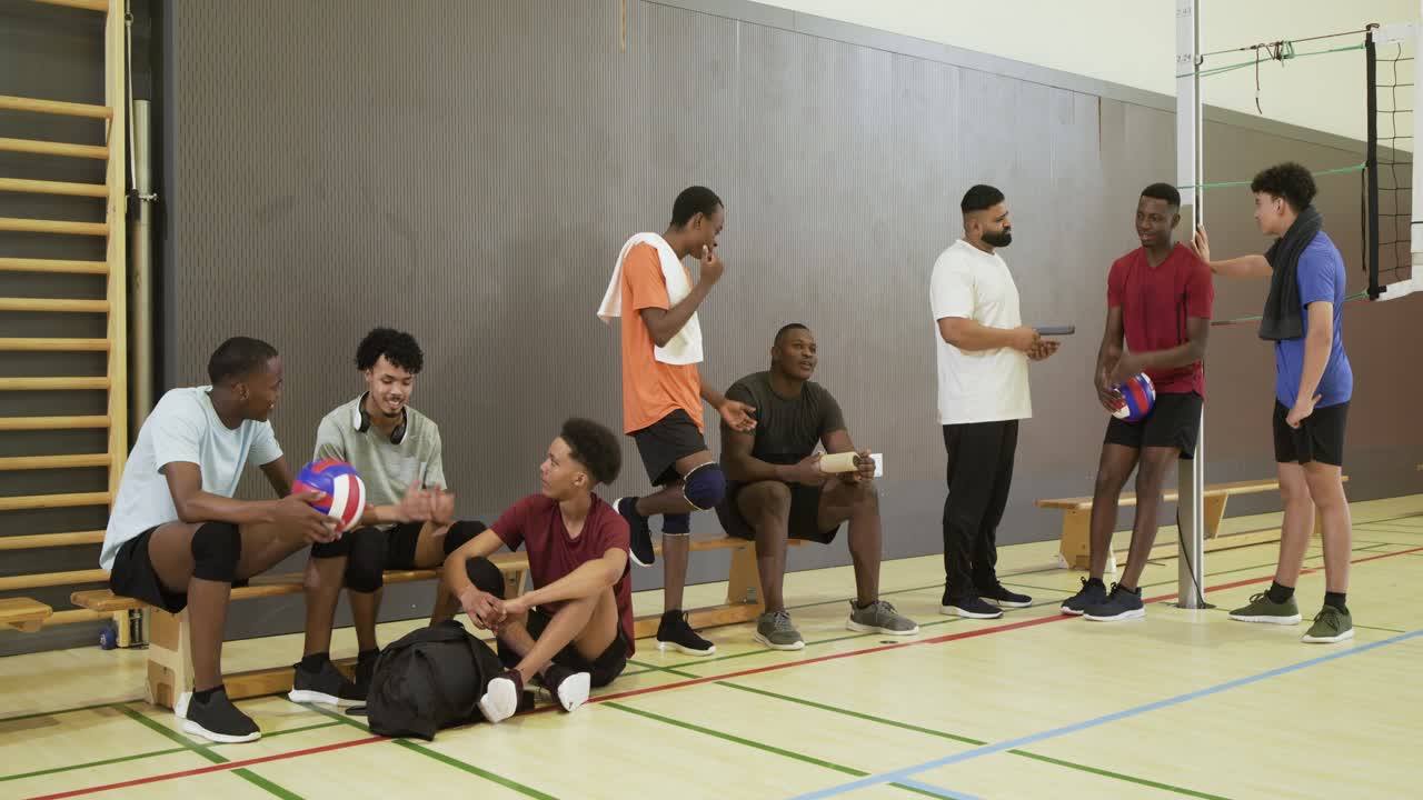 Diverses Männerteam sitzt während einer Auszeit auf der Bank in der Schulsporthalle, passt einen Volleyball und überprüft das Handy