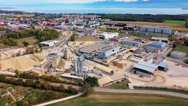 A drone pans then orbits La Ballastiere in Gland, Vaud, Switzerland, showing silos, conveyors, sand and gravel stockpiles, with the town, lake and mountains behind.