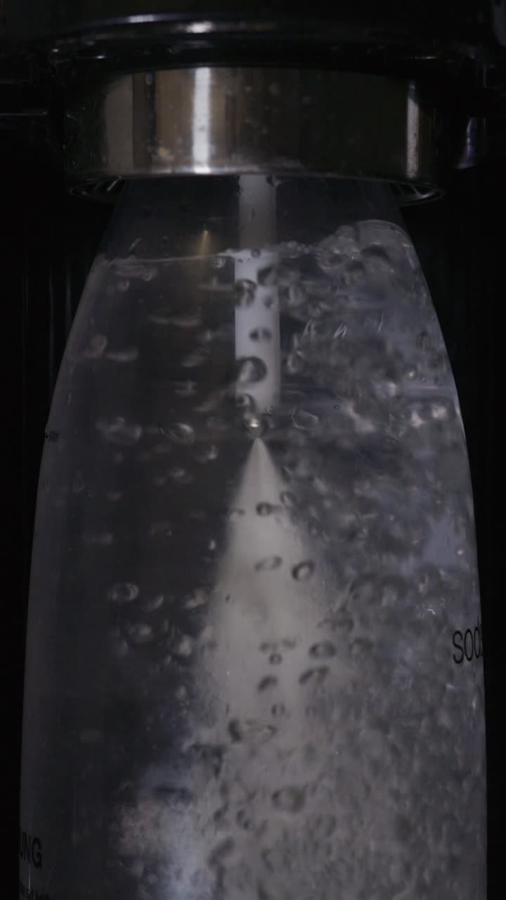 Pionowy widok z bliska urządzenia do karbonizacji, które wstrzykuje CO₂ do plastikowej butelki wypełnionej wodą. Unoszące się bąbelki i musujący spray tworzą dynamiczną teksturę wewnątrz pojemnika.