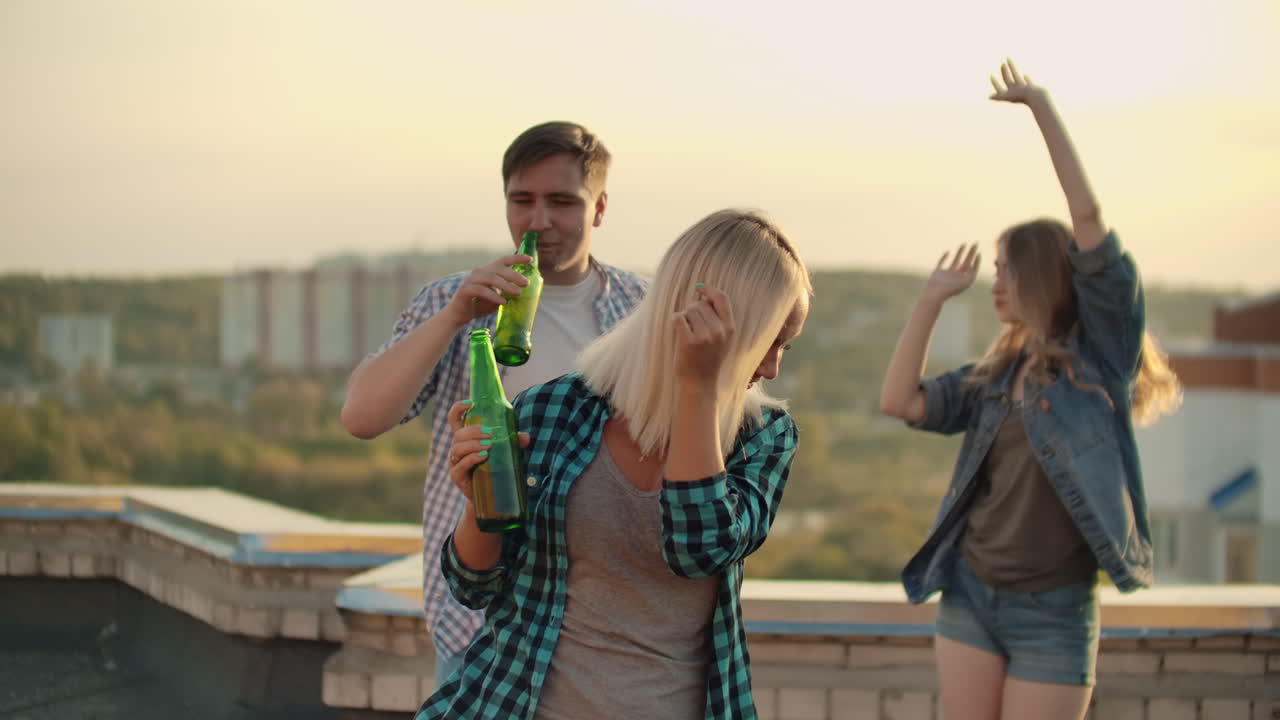 The American blonde woman is dancing on the roof with her friends on the party. She smiles and enjoys the time in shorts and she is dancing with a beer in a gray tank top and a dark green plaid shirt.