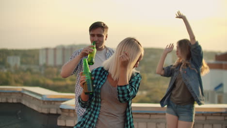 the american blonde woman is dancing on the roof with her friends on the party. she smiles and enjoys the time in shorts and she is dancing with a beer in a gray tank top and a dark green plaid shirt.