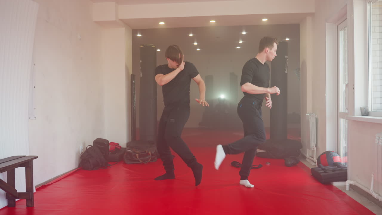 Fighters practicing martial arts in gym, one in defensive stance with hands up while other faces him sparring, red mats on floor, punching bags in background, focused training atmosphere under bright lights