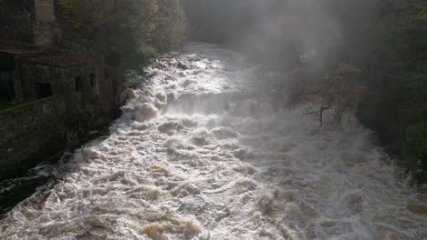 Drohnenaufnahmen-Fliegen-In-Der-Nähe-Eines-Schnell-Fließenden-Flusses-Mit-Blick-Auf-Einen-Wasserfall,-Der-Im-Herbst-Von-Einem-Wald-Aus-Laubbäumen-Umgeben-Ist