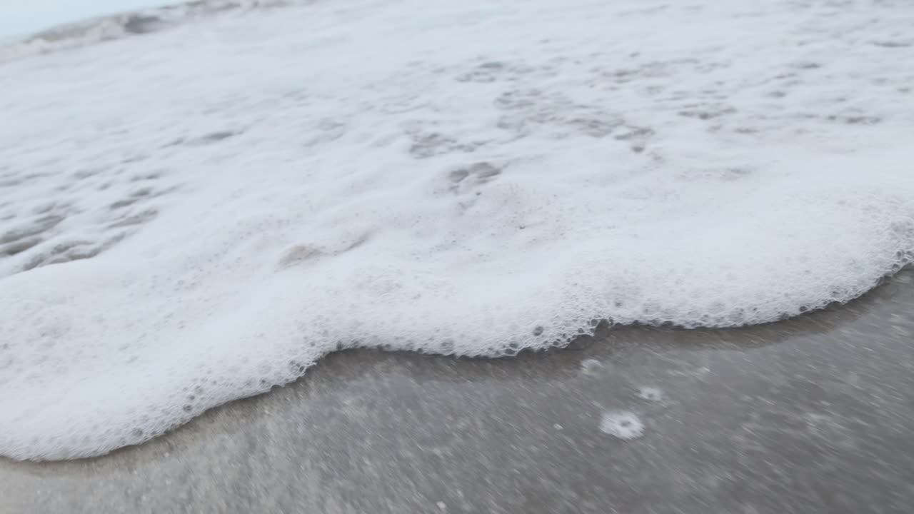 Foamy sea water washing up on a sandy beach slow motion