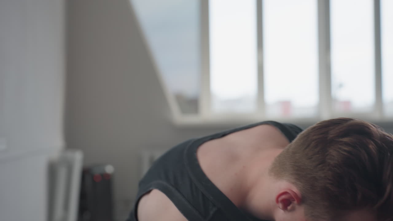 Young man in black tank top leans over navy suitcase by sunlit apartment window, retrieves personal items and places them on windowsill, preparing belongings in minimalist modern room interior