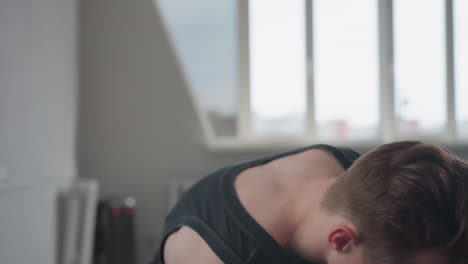 young man in black tank top leans over navy suitcase by sunlit apartment window, retrieves personal items and places them on windowsill, preparing belongings in minimalist modern room interior