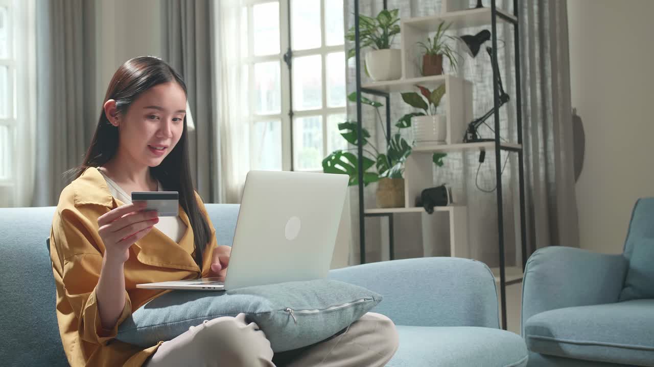Asian Woman Sitting On Couch In Living Room Holding Credit Card And Celebrating While Using Laptop Computer Shopping Online