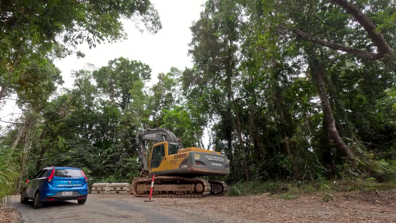 Blue car drives through lush rainforest, encountering roadwork and excavator under overcast natural lighting