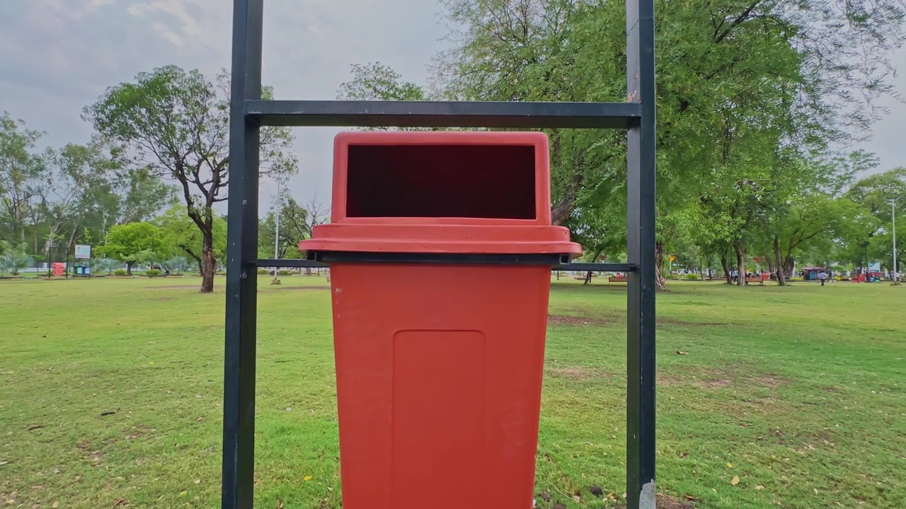 Acercamiento suave de cámara hacia un moderno contenedor de basura público instalado en un parque urbano ajardinado, destacando la infraestructura de limpieza urbana y el entorno verde.