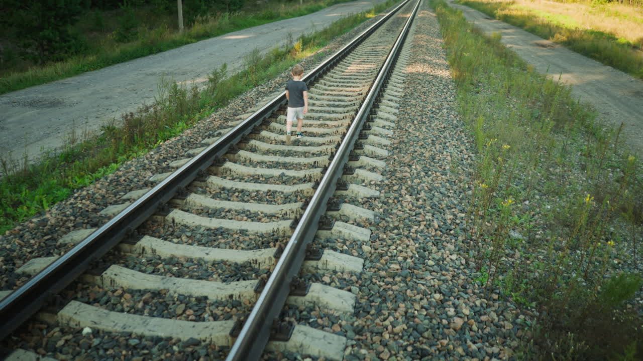 rear view of young boy in gray t-shirt and light shorts walking confidently along rail track through countryside, with dirt roads and overgrown greenery on both sides