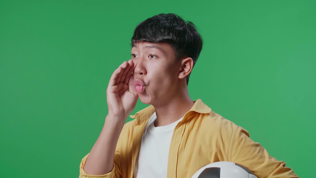 초록색 스크린 배경에서 축구 선수를 응원하는 동안 패자들을 조롱하는 공을 들고 있는 아시아 소년의 가까운 측면 뷰