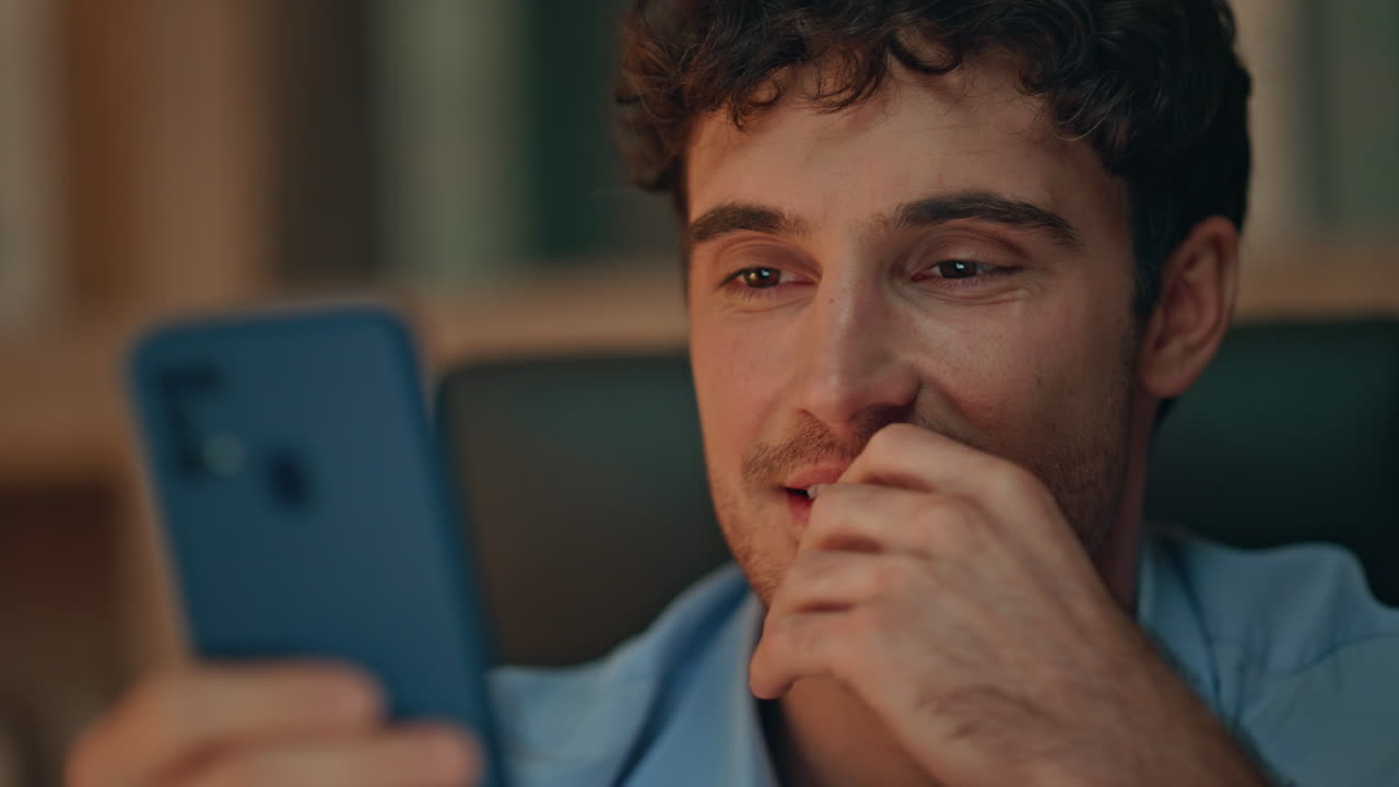 Freelancer despreocupado navegando por conteúdo de vídeo no celular em casa à noite, em close-up.