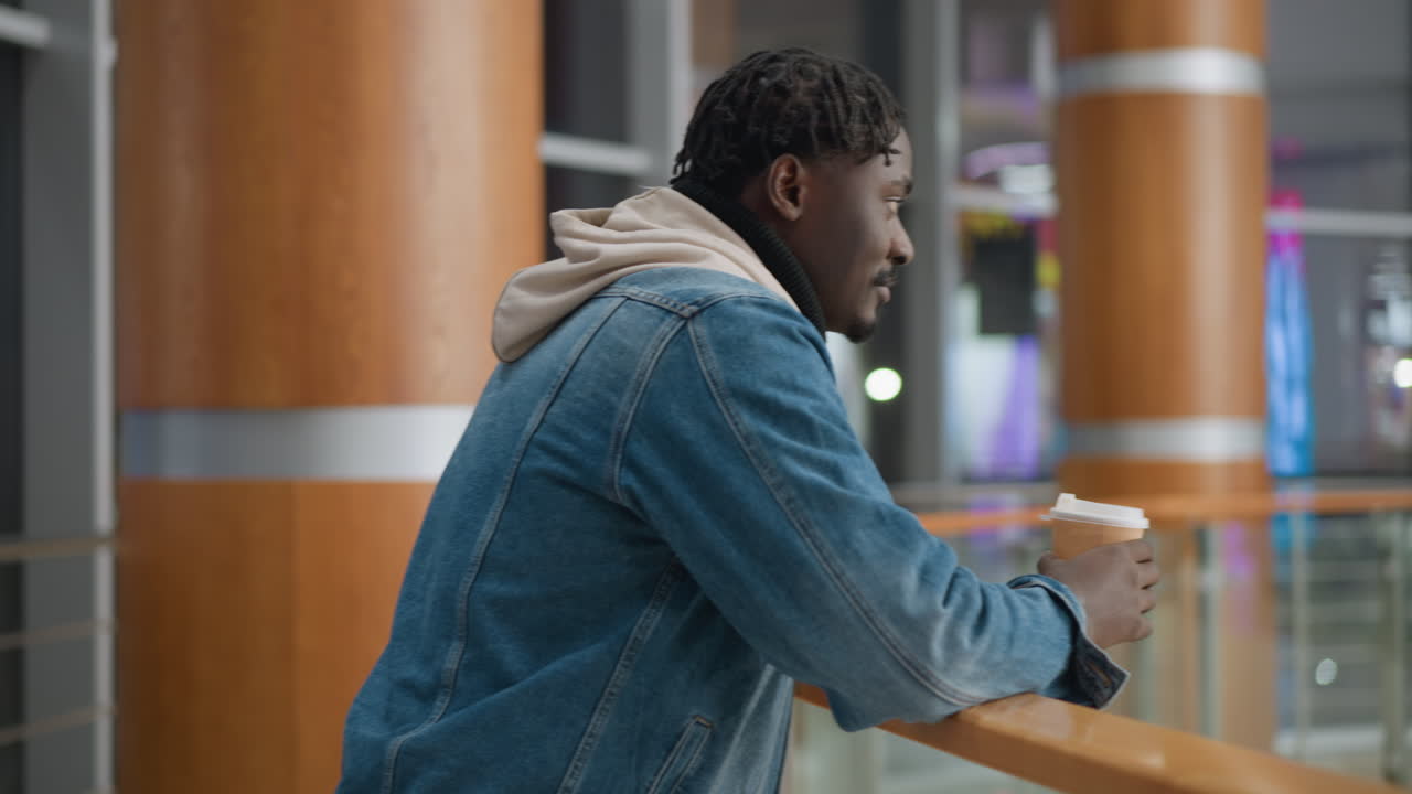 Young man in casual denim jacket and hoodie holding paper cup, standing by glass railing in modern indoor space, gazing thoughtfully and sipping hot beverage, enjoying peaceful moment