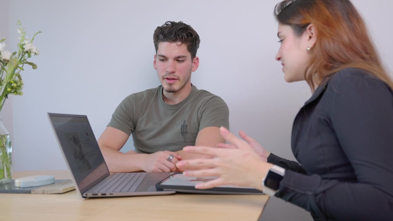 Dolly in zoom shot of a brunette woman talking to a Caucasian male in front of a touchscreen laptop discussing a tattoo project while sitting on a table with flowers in a brightly lit scene