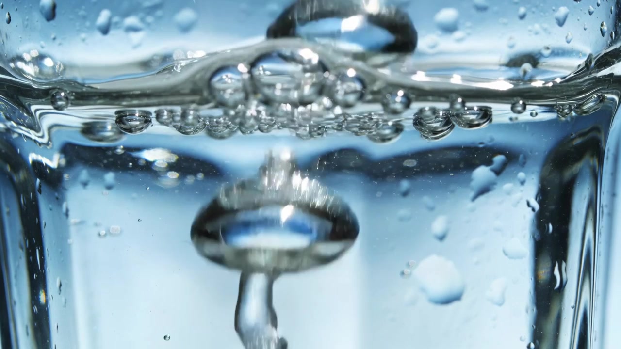 Pouring water stream forming column inside clear glass in studio to fill glass, large bubble rising
