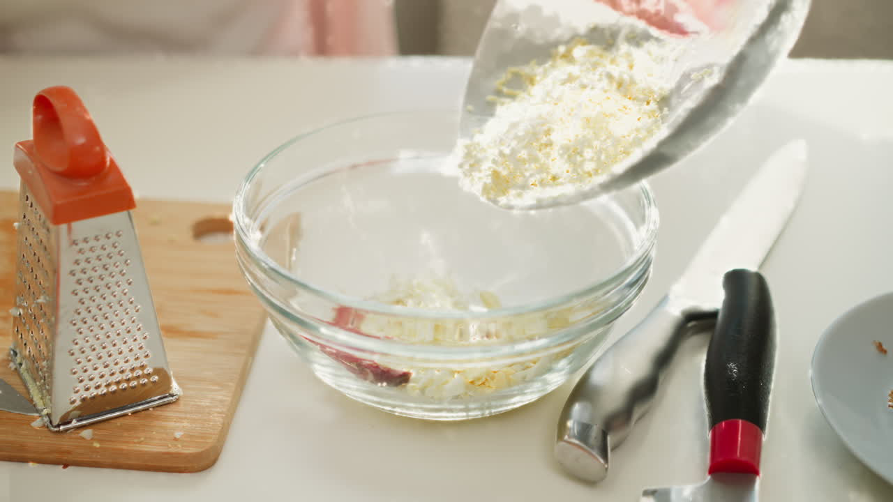 Chef hand carefully pouring grated boiled egg from plate into transparent bowl containing sliced egg and sausage on kitchen table, preparing ingredients for delicious homemade meal under bright light