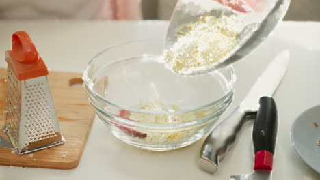 chef hand carefully pouring grated boiled egg from plate into transparent bowl containing sliced egg and sausage on kitchen table, preparing ingredients for delicious homemade meal under bright light