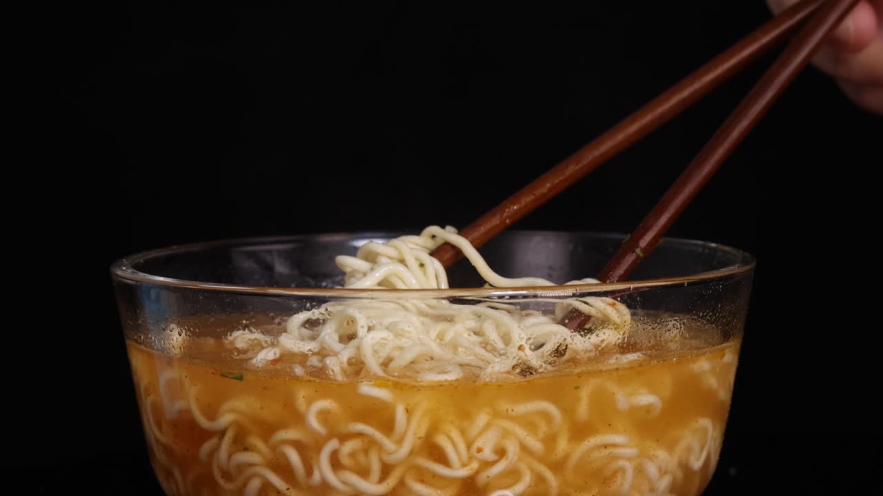 A hand skillfully lifts cooked instant noodles from a glass bowl of broth using wooden chopsticks, under bright studio lighting with a black background