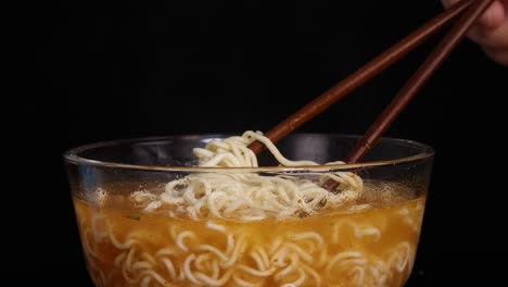 a hand skillfully lifts cooked instant noodles from a glass bowl of broth using wooden chopsticks, under bright studio lighting with a black background
