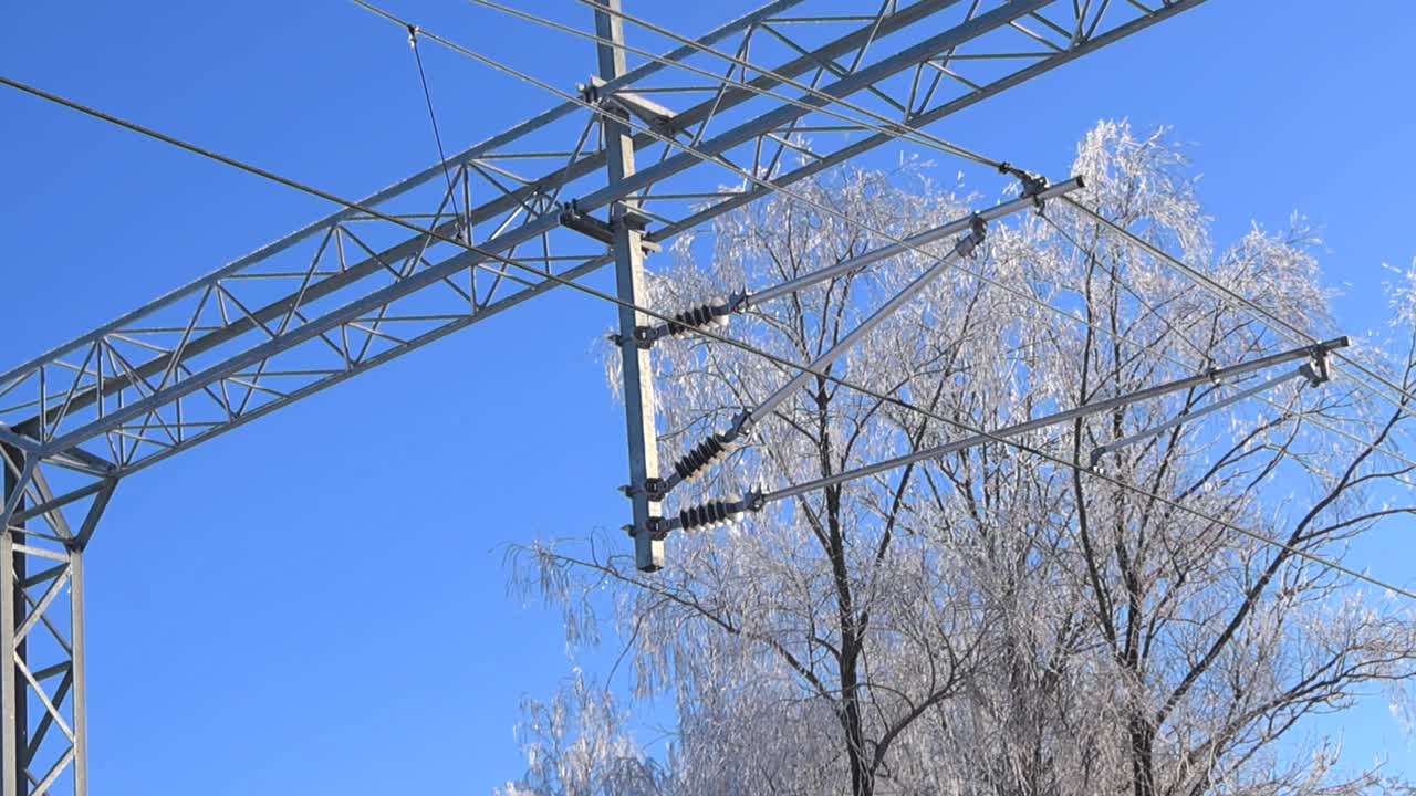Nedefra og op-udsigt over frossent jernbaneelektrificeringsudstyr og kabler, der glitrer mod blå himmel og snedækkede træer