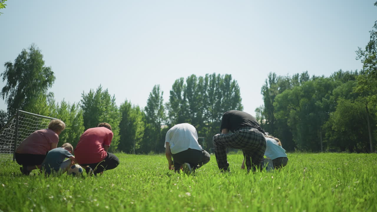 Back view of a family in groups of three tying their shoelaces on a sunny day, with a small goalpost visible nearby