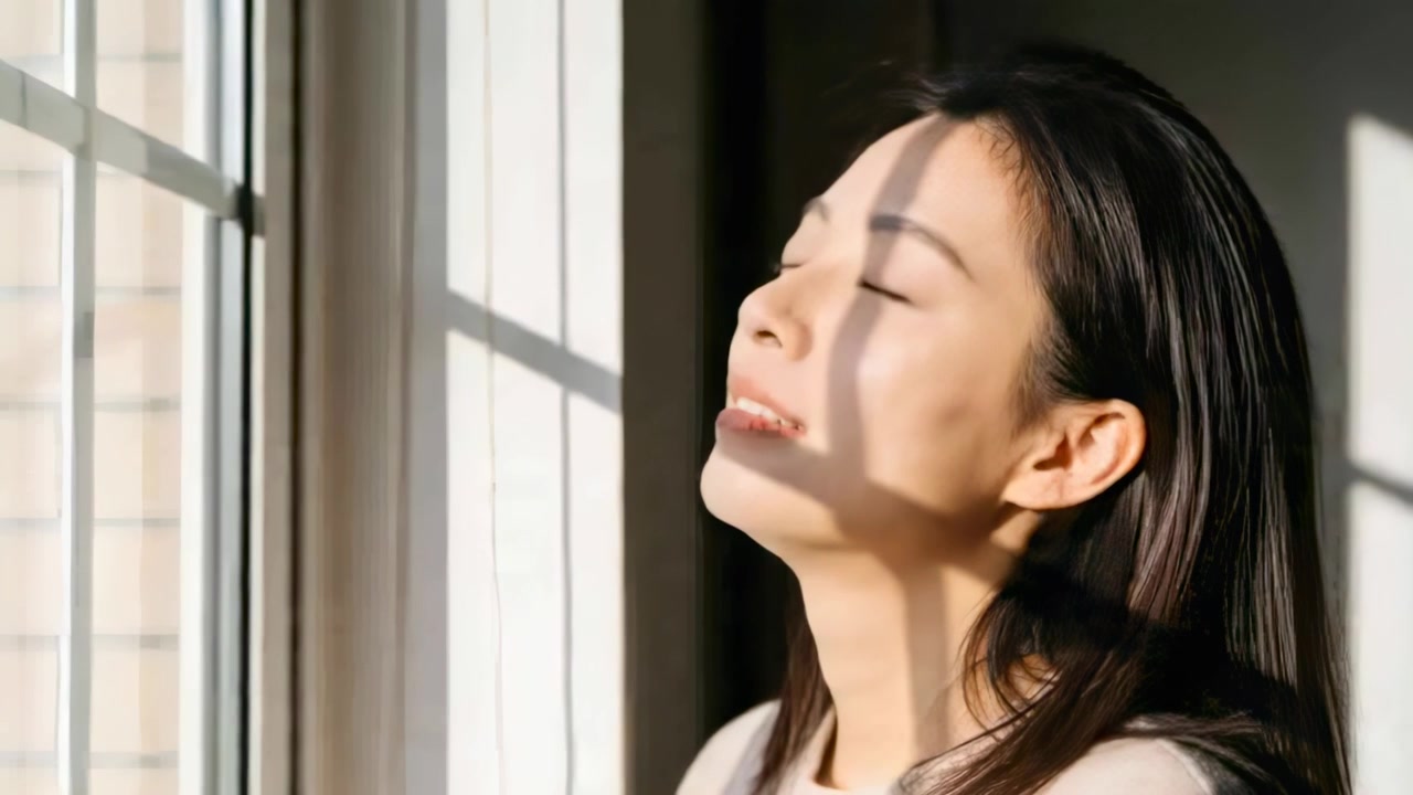 自宅でセーターを着たアジア人女性が、太陽に向かって顎を上げ、格子状の影が落ちる中で日光浴をしている。コピースペースあり。