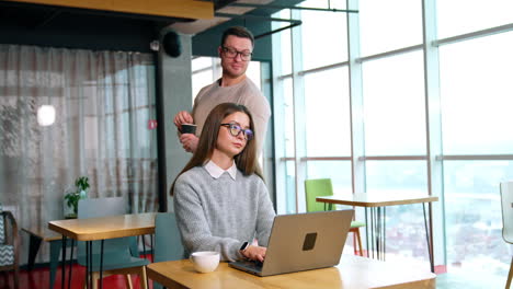long-haired female working at her laptop in the office. man stirring sugar in the cup comes up behind peeping at colleagues computer.