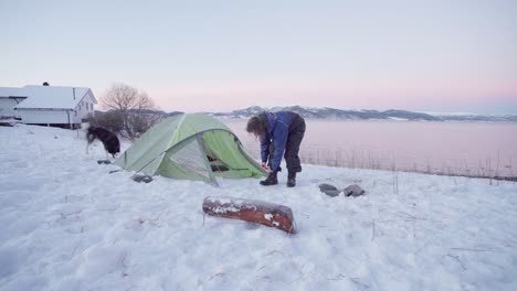 man tying shoelace of black leather boots in snowy campground by the lakeshore with playful alaskan malamute running around at sunset