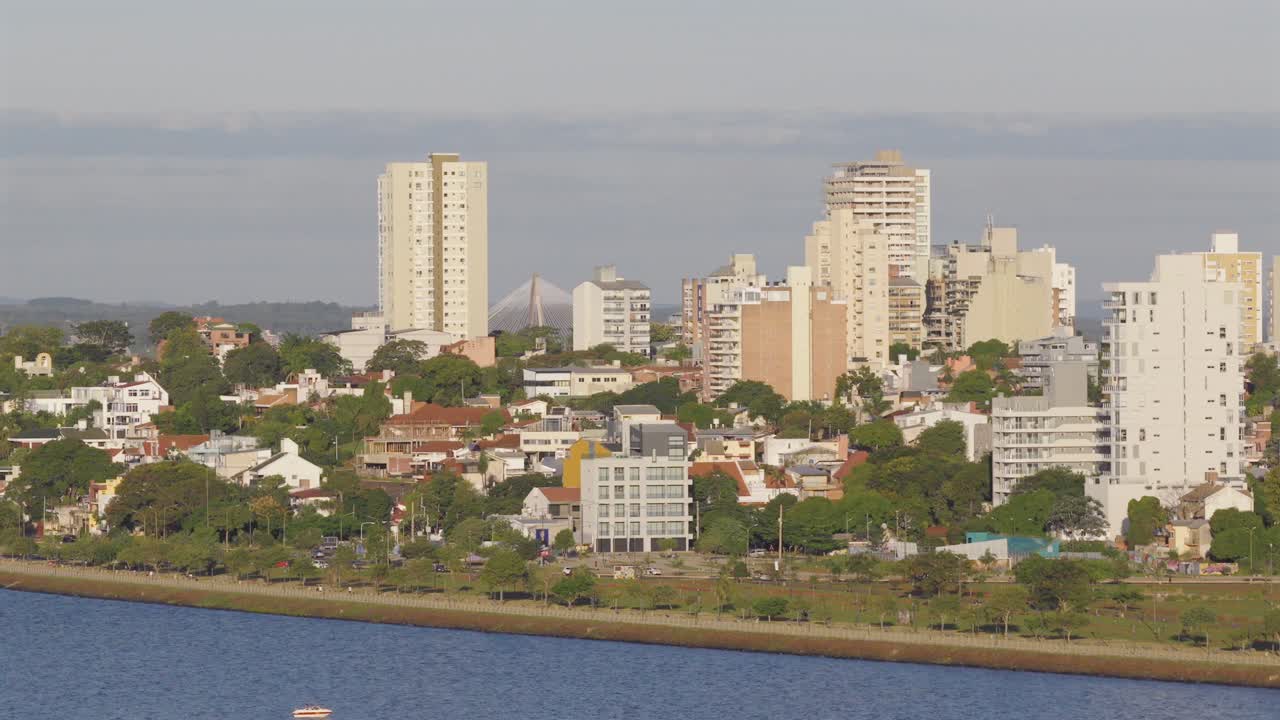 Drone shot of Posadas city skyline in Misiones, Argentina, showing tall residential and commercial buildings, urban green areas, riverside promenade, and the Paraná River on a sunny day, real time.