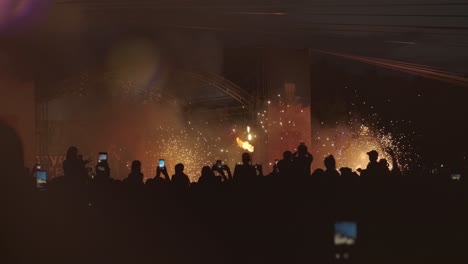 a large group of people filming fireworks and a fire show on their mobile phones. vibrant flames and colorful fireworks light up the night sky, creating an electrifying atmosphere