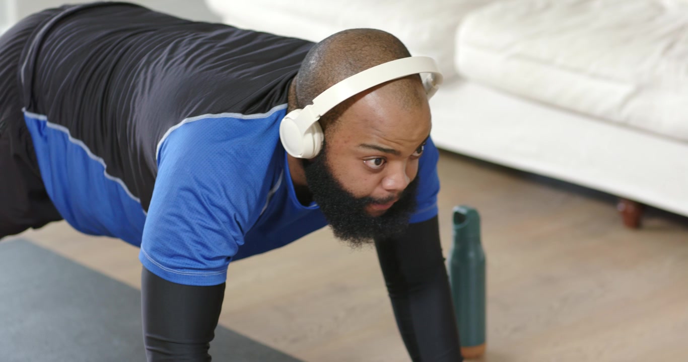 Uomo afroamericano adulto con una maglietta blu che indossa una cuffia, esegue sollevamenti delle ginocchia su un tappetino seguendo un audio.