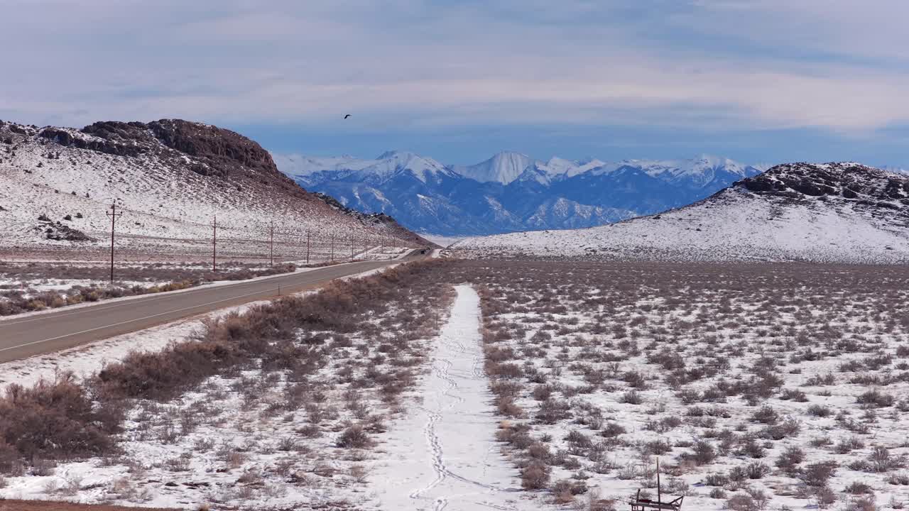 Un’ampia e spettacolare inquadratura di una strada innevata che conduce verso l'imponente catena montuosa Sangre de Cristo. Un corvo (o cornacchia) vola in primo piano sopra il paesaggio ad alta quota.