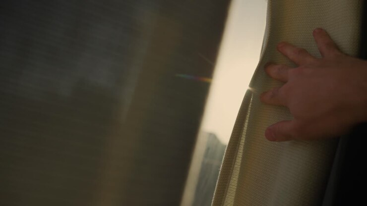 Hand reaching towards window curtain to reveal sunlight shining from outside. Person opening and drawing shade to let sun shine inside dark apartment or house. Hope and positive energy green solutions