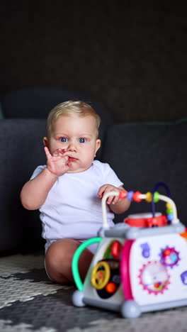 beautiful caucasian blond toddler sits on the floor near his toy. kid looks surprised ahead and chews his finger. vertical video.