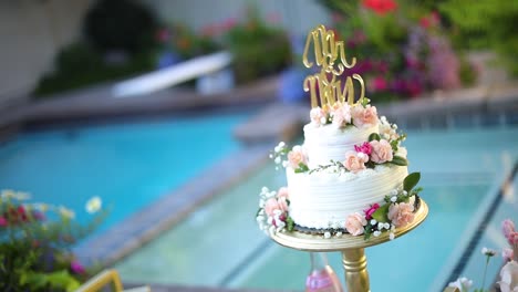 shot of a beautifully decorated floral wedding cake sitting on a pedestal right next to a pool during a summer wedding reception