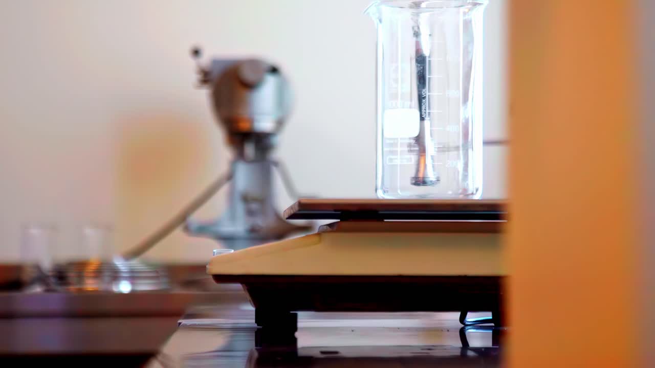 Un primo piano di una bilancia analitica di precisione da laboratorio e un alto becher di vetro su un banco di legno, con attrezzature vintage sfocate sullo sfondo