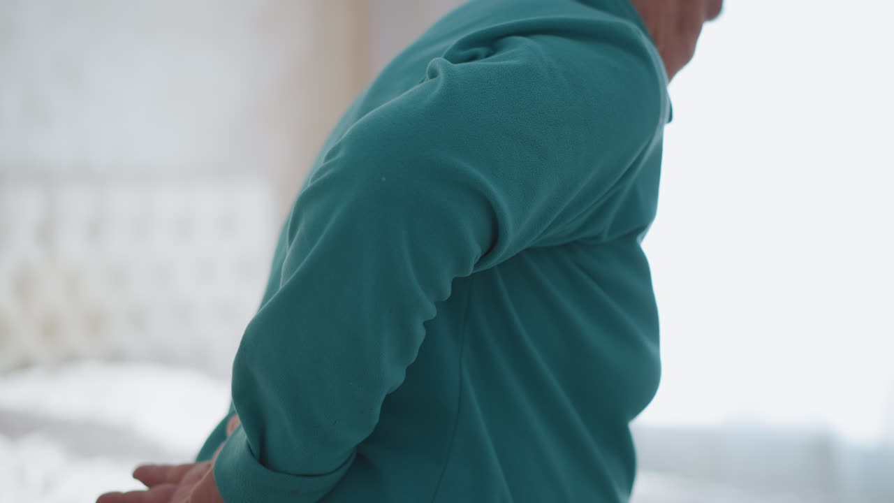 Elderly woman in green jacket, bent over slightly with a hand placed on her lower back, appearing to stretch or ease discomfort, sitting indoors in a bright room, serene atmosphere