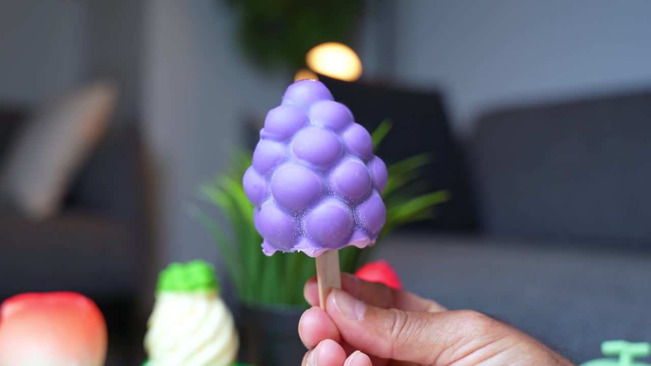 Colorful close-up of grape-shaped purple ice cream bar held by hand with soft background. Great for summer, kids’ treats, dessert ads, and playful food visuals.
