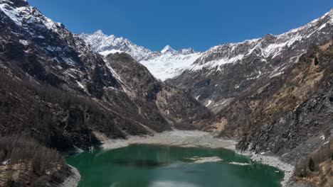 aerial slow dolly forward shot of a green alpine lake nestled at the base of a ring of snow capped mountains