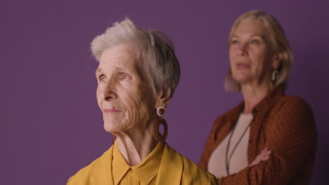 blonde senior woman with short hair wearing mustard colored shirt and jacket and earrings, posing with blurred mature woman on purple background 1