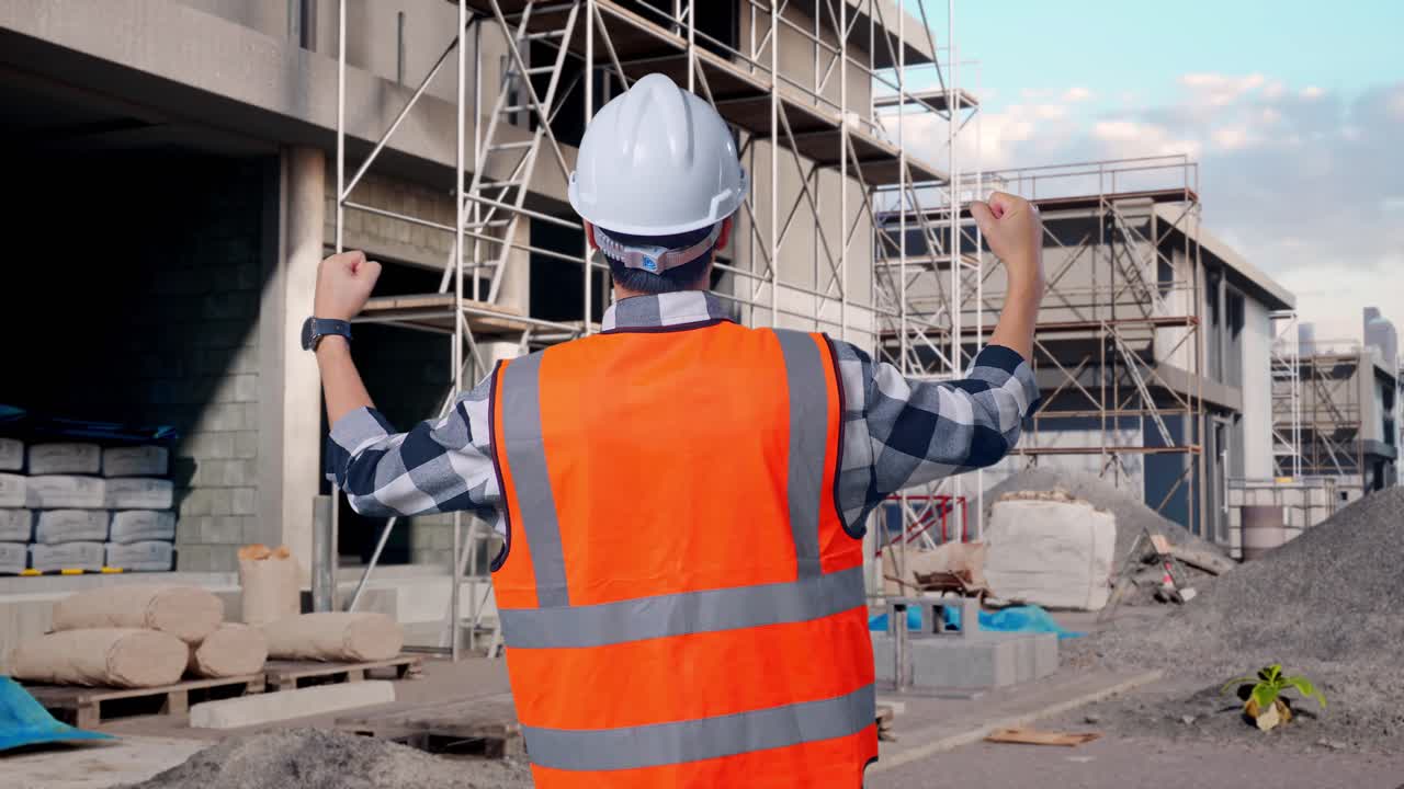 Back View Of A Male Engineer With Safety Helmet Raising His Hands Celebrating While Working At Building & Construction Site in Progress to New House