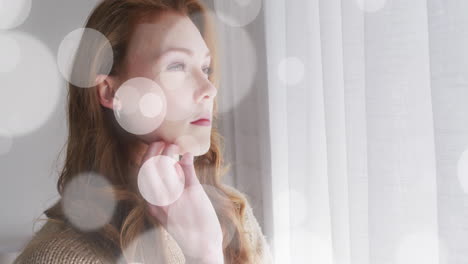 Animation-of-light-spots-over-caucasian-woman-looking-through-window