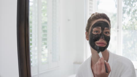 Portrait-of-biracial-woman-applying-beauty-face-mask-in-bathroom,-slow-motion