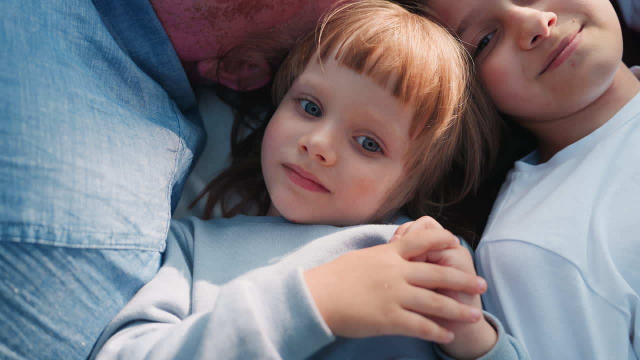 Ingrandisci una bambina sdraiata tra la sorella e il papà durante un momento di relax all'aperto, che sorride dolcemente sotto la calda luce del sole, catturando il pacifico legame dell'infanzia e la calma connessione familiare