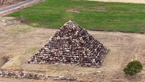 drone camera smoothly circles a large granite rock pyramid in a dry, grassy field near ballandean, queensland, under natural daylight with steady movement