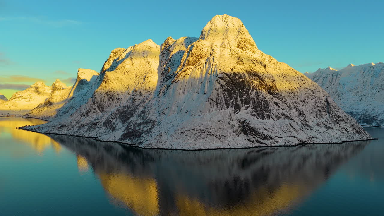 Cinematic aerial view of snowcovered peaks reflecting in calm fjord waters during golden hour in Lofoten, Norway. Great for travel and nature documentaries