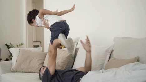 Joyful-dad-lying-on-couch,-holding-and-lifting-son