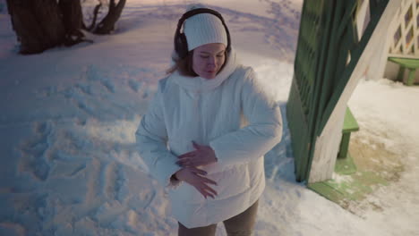 female artist wearing headphones sways gently in snowy outdoor environment under bare winter trees with rhythmic body movement and soft evening light highlighting flowing coat and crisp winter air