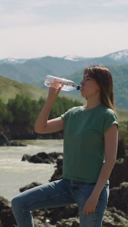 thirsty woman drinks mineral water on rocky bank of mountain river on hot summer day. prevention overheating at hike. healthy lifestyle