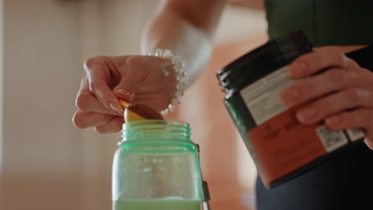 Donna che fa fitness in cucina a casa che mette la polvere nello shaker, primo piano delle mani che aprono il contenitore e aggiungono un misurino, etichetta testurizzata, concentrazione silenziosa, preparazione per l'allenamento della forza e il recupero